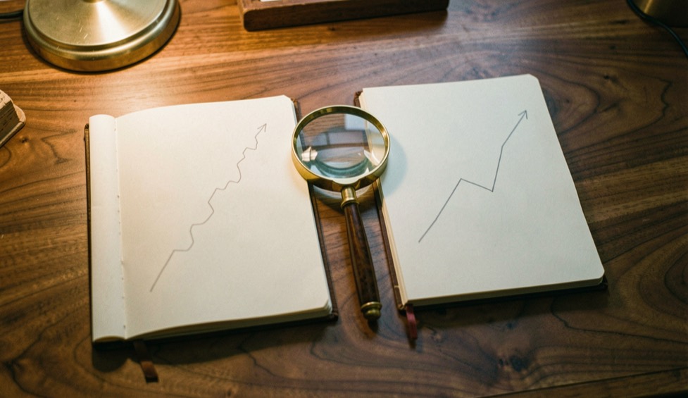 Two open paper notebooks side by side on a walnut desk, each with a pencilled line climbing at a different pace, a brass magnifying glass resting between them, warm lamp light in the evening